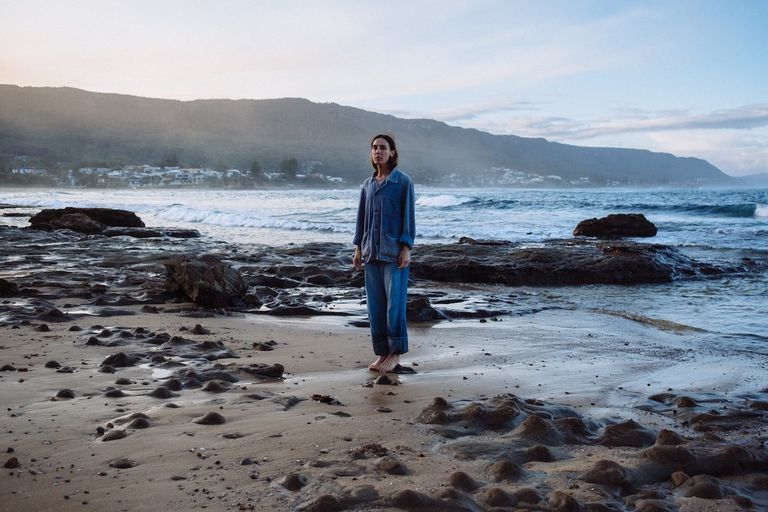 Musician Holly Throsby standing barefoot on a rocky beach wearing denim, with ocean waves and a coastal mountain in the background